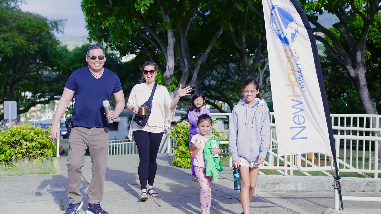 A family walking in under the monkeypod trees past a New Hope Hawaii Kai flag, mom waving