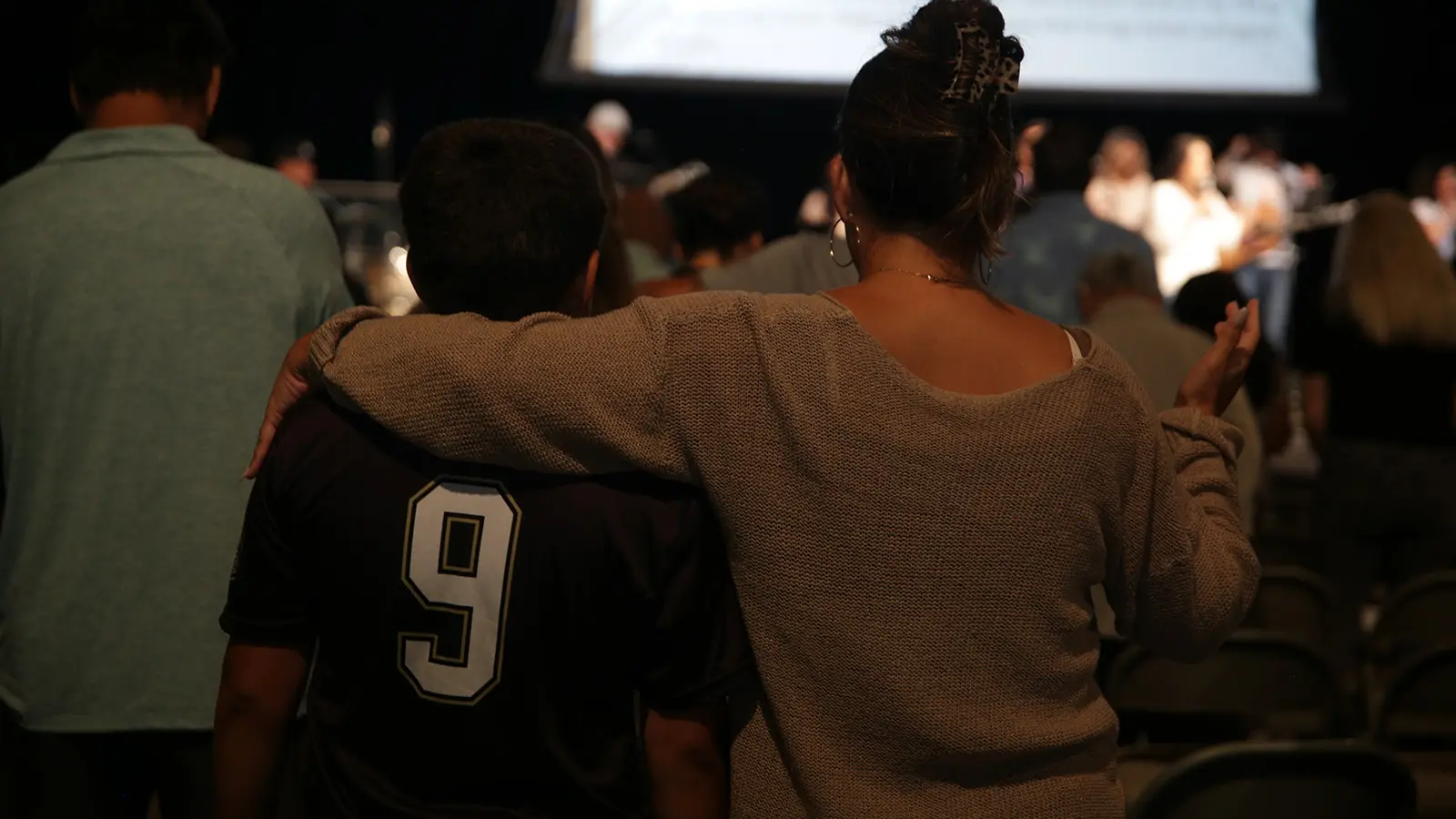 Two people from behind, one with an arm around the other's shoulder, during worship