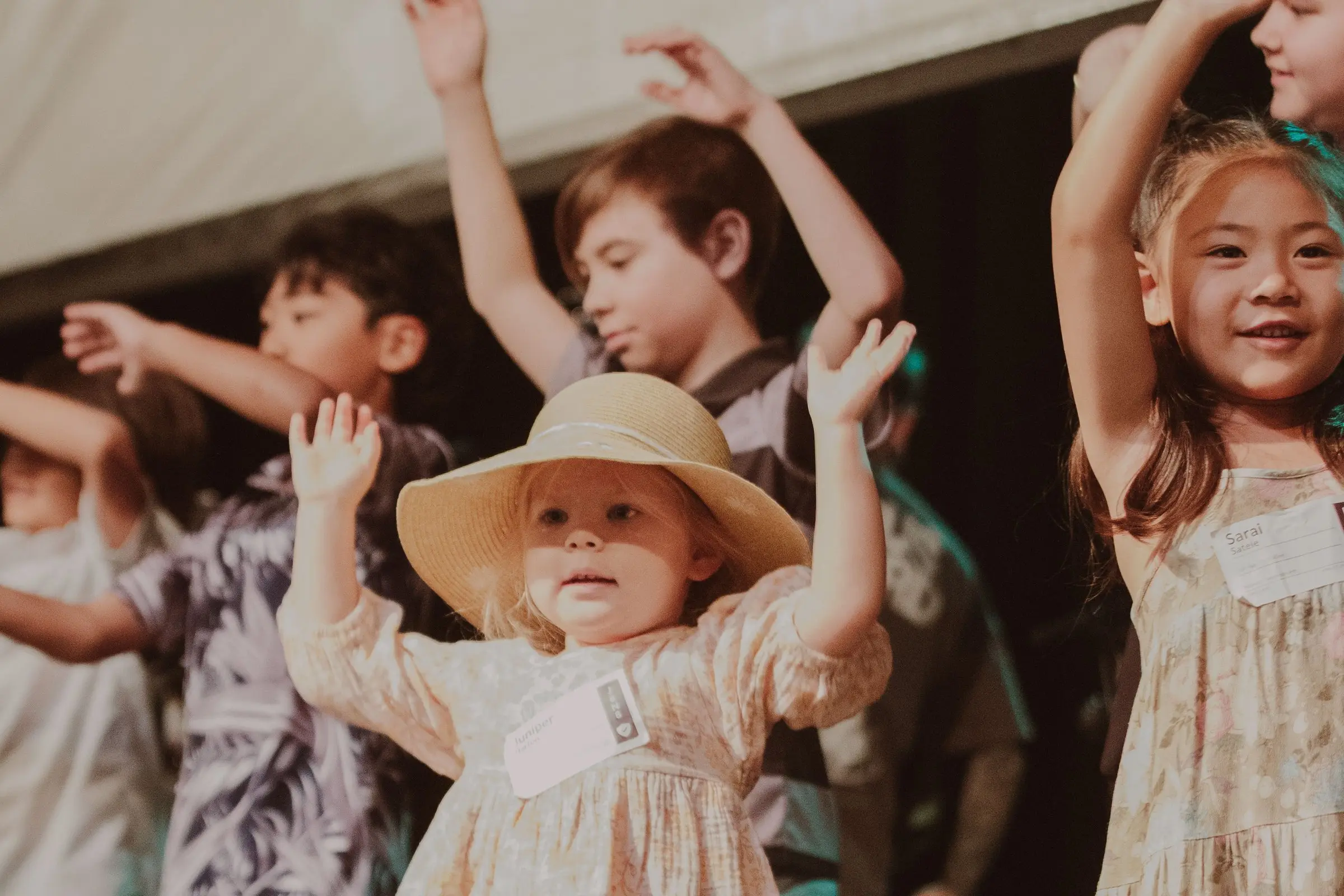 Kids with their hands up singing on stage on Easter Sunday, a toddler in a sun hat in front
