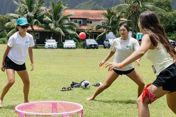 Teens playing spikeball on a field with palm trees and the Koolau cliffs behind